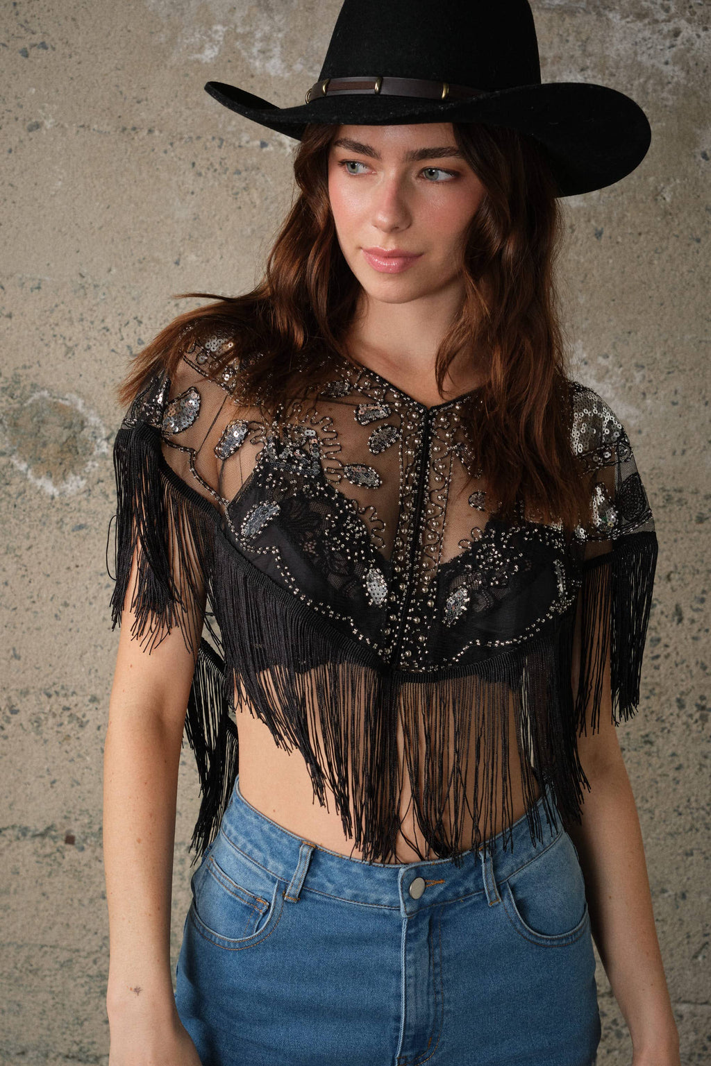 Sheer black mesh cape top with sequin and bead details, fringe trim, and open front, styled over high-waisted denim.