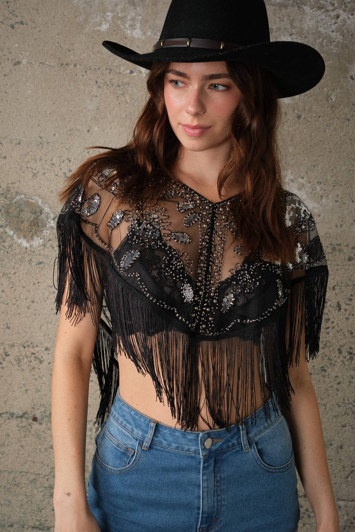Sheer black mesh cape top with sequin and bead details, fringe trim, and open front, styled over high-waisted denim.