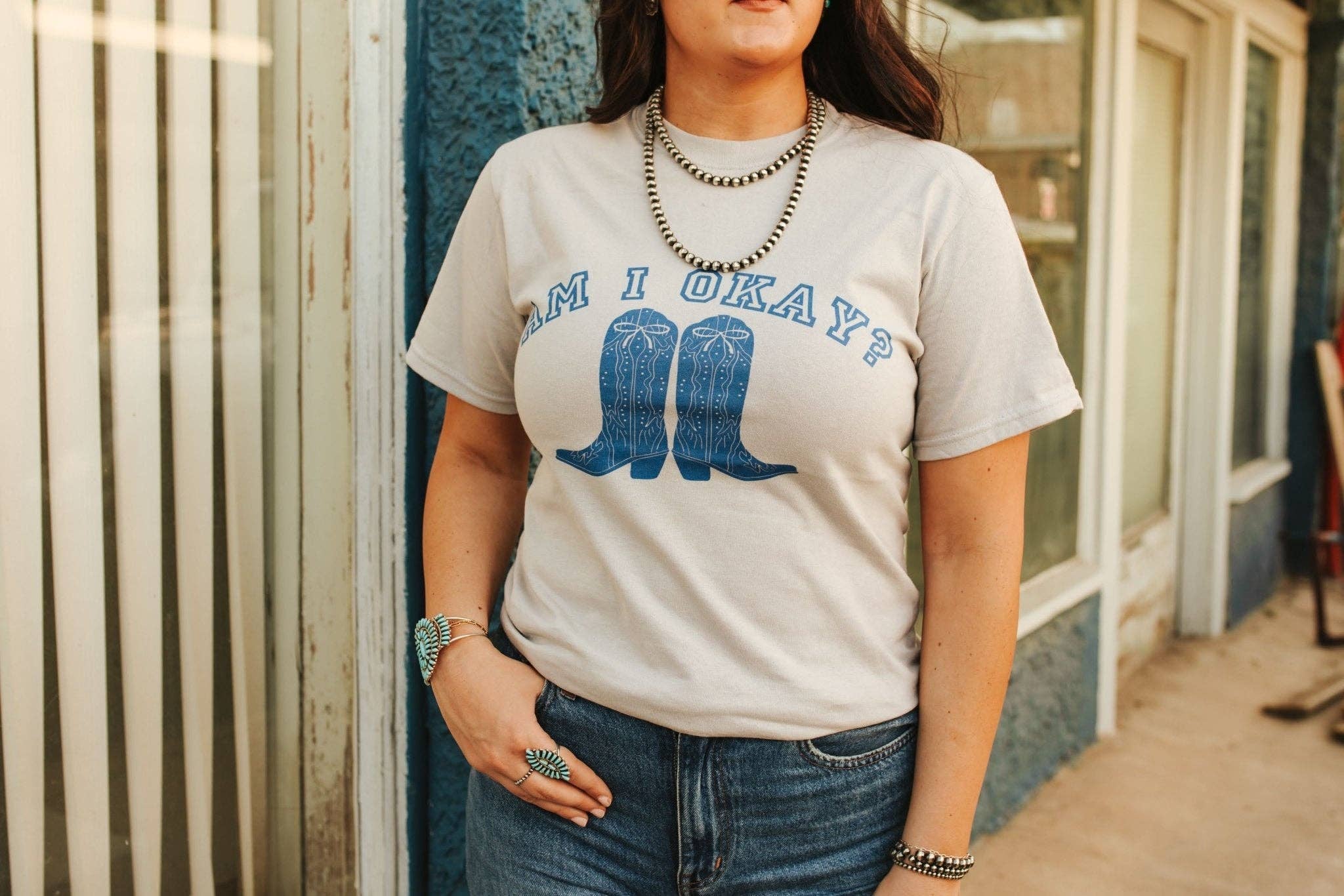 Woman wearing unisex beige Am I Okay Graphic Tee with blue cowboy boots print and jeans standing outdoors