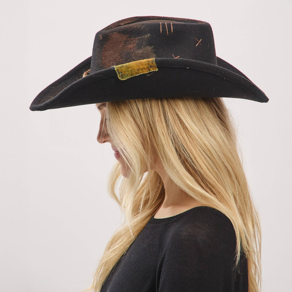 Rustic shapeable cowgirl fedora hat in black with distressed fabric band, worn sideways by a model with long blonde hair.