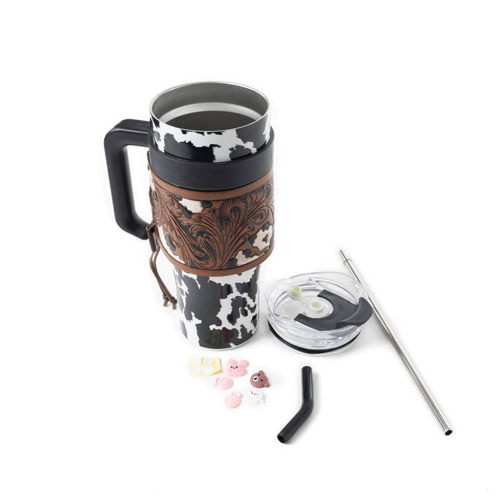 Black and white cowhide print tumbler with chocolate brown tooled leather wrap, ergonomic handle, and spill-resistant lid.