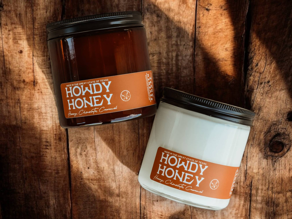 Howdy Honey caramel, vanilla, and honey scented candle in clear and amber jars on wooden surface