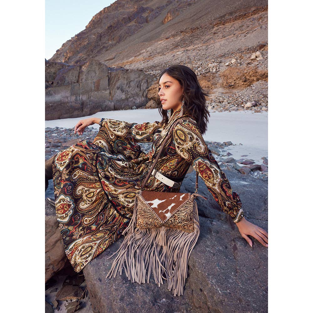 A model sits on rocks wearing a paisley patterned dress, showcasing the Harappa Leather & Hairon Bag with fringe details.