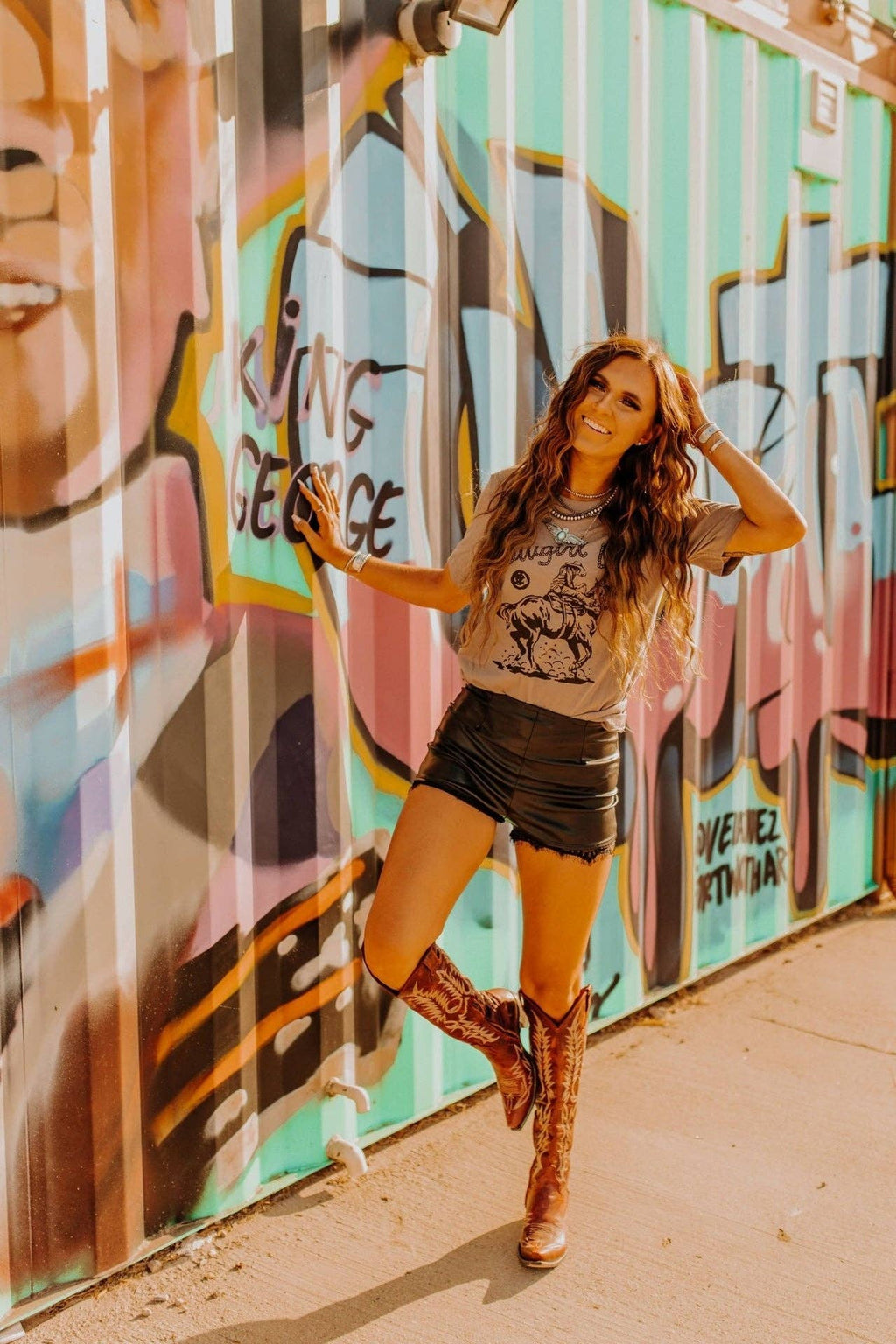 Woman wearing Cowgirl Up Graphic Tee with cowboy boots and shorts standing by colorful graffiti wall