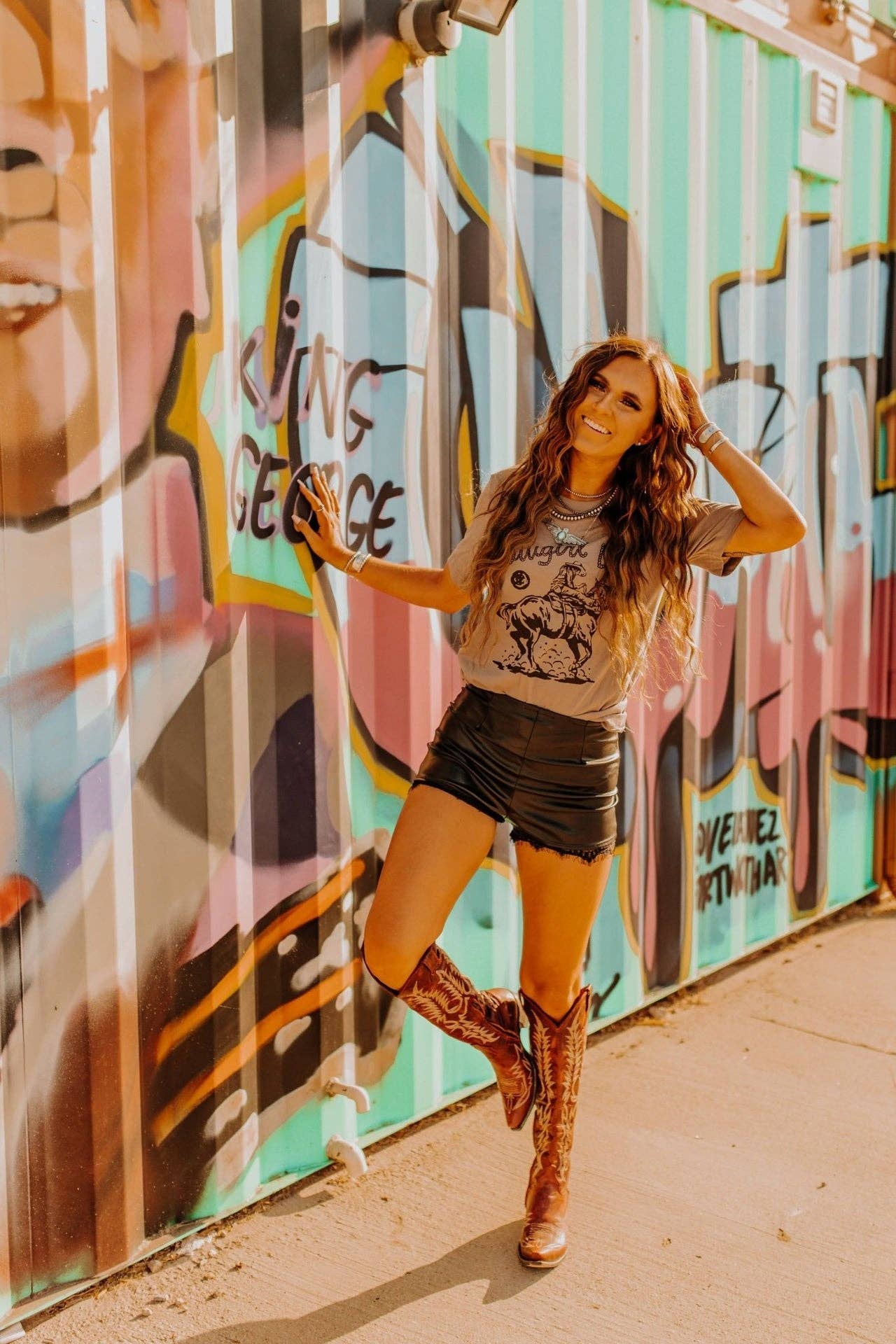 Woman wearing Cowgirl Up Graphic Tee with cowboy boots and shorts standing by colorful graffiti wall