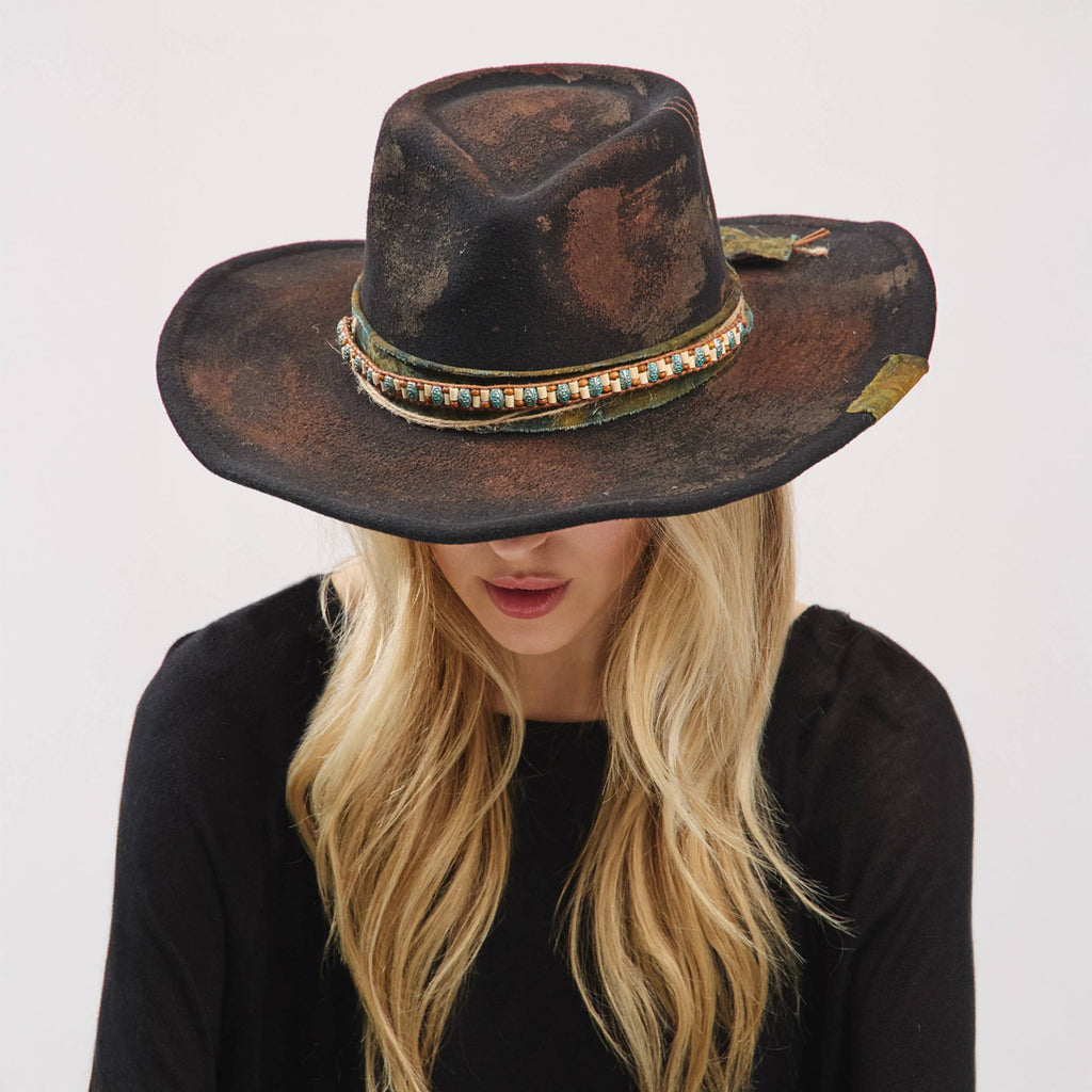 Distressed black cowgirl fedora with a colorful fabric band, worn low, showcasing a rustic and vintage style.