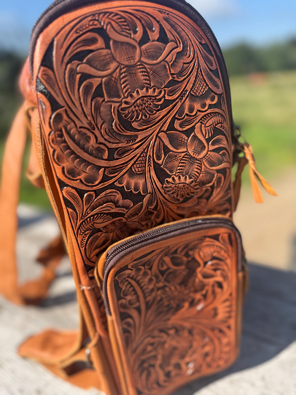 Genuine leather brown backpack with intricate tooled floral patterns, featuring adjustable straps and multiple zippered pockets.